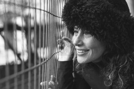 Middle aged charming lady with Russian fur hat holding to fence with smile observing wildlife at zoo park. Fall fashion sales concept. Black and white photography