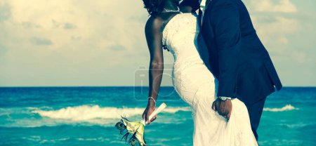 Photo for Passionate Afro-American couple kissing on wedding day by the sea. Bride holding bouquet and groom grabbing her leg on the beach. Ceremony union, Valentine Day, love concept. Vintage effect - Royalty Free Image