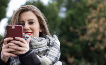 Photo for Close up of blonde girl holding red mobile phone on cold autumn day at park. Young woman, cellphone addiction, online dating app, communication, new technologies, trendy concepts - Royalty Free Image