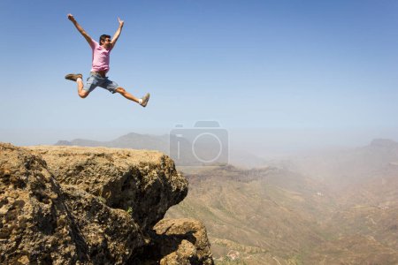 Mutiger Mann im rosafarbenen T-Shirt, der hoch oben auf einer Felskante im Naturpark Roque Nublo auf Gran Canaria springt. Junger Bergsteiger feiert auf dem Gipfel. Risiko, Gefahr, Wagnis, Furchtlosigkeit, Abenteurer, Erfolgskonzepte