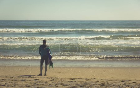 Foto de Espalda de surfista en traje de neopreno con tabla corporal y aletas en la mano de pie en la orilla observando olas en día soleado en la playa de La Serena, Chile. Efecto vintage - Imagen libre de derechos