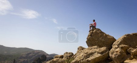 Foto de Hombre solitario sentado en la cima de la roca en un día soleado en el Parque Nacional Roque Nublo, Gran Canaria. Excursionista observando paisaje natural panorámico de montaña en Canarias, España. Explorador, conceptos visionarios - Imagen libre de derechos