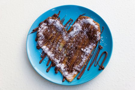 Foto de Panqueque en forma de corazón con crema de chocolate y coco rallado sobre plato azul y fondo blanco. Delicioso postre dulce - Imagen libre de derechos