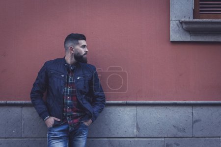 Foto de Joven hipster parado en la calle mirando hacia un lado. Modelo masculino de moda con jeans azules, chaqueta y camisa de damas apoyadas en la pared. Efecto oscuro - Imagen libre de derechos
