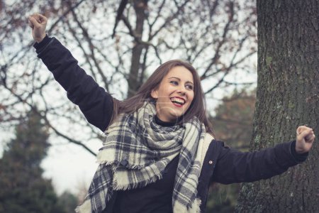 Photo for Joyful brunette girl smiling with arms up in the park on cloudy autumn season day. Celebration concept. Pretty young woman happy as if having won the lottery - Royalty Free Image