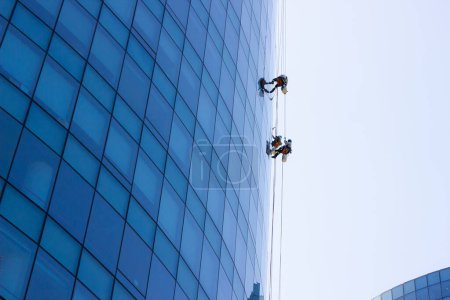 Téléchargez les photos : Ouvriers de nettoyage de fenêtres suspendus à l'extérieur du bâtiment de bureaux en verre bleu. Travail risqué, concepts de travail dangereux - en image libre de droit