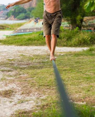 Téléchargez les photos : Gros plan sur les jambes de l'homme en short gardant l'équilibre tout en marchant sur slackline à la plage dans l'île de Koh Phangan, Thaïlande. Promenez-vous, concepts d'équilibre - en image libre de droit