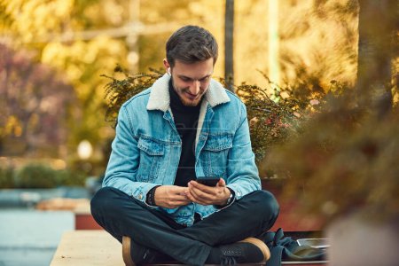 Ein Mann, der auf einer Bank in der Innenstadt sitzt und sein Smartphone und drahtlose Kopfhörer für einen Anruf benutzt, zeigt die Bequemlichkeit der Technologie in der modernen Kommunikation und die Fähigkeit, Multitasking zu betreiben, während