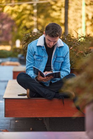 Téléchargez les photos : Un homme assis sur un banc dans le centre-ville lisant un livre tout en portant un casque sans fil, démontrant la puissance de la littérature et la capacité de déconnecter les distractions dans un environnement occupé en utilisant - en image libre de droit