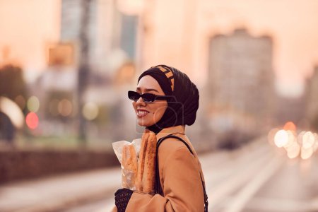 Foto de Una mujer hiyab, vestida con un lujoso atuendo vintage francés, mientras camina por las calles de la ciudad al atardecer, llevando pan, un ramo de flores y periódicos en su mano, evocando una cautivadora mezcla - Imagen libre de derechos