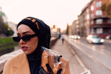 Foto de Una mujer hiyab, vestida con un lujoso atuendo vintage francés, mientras camina por las calles de la ciudad al atardecer, llevando pan, un ramo de flores y periódicos en su mano, evocando una cautivadora mezcla - Imagen libre de derechos