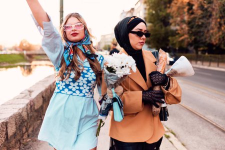 Photo for Couple woman one wearing a hijab and a modern yet traditional dress, and the other in a blue dress and scarf, walking together through the city at sunset. One carries a bouquet and bread, while the - Royalty Free Image