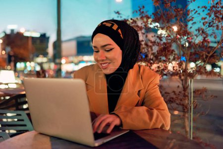 Photo for In the enchanting ambiance of a nocturnal cityscape, a hijab-clad girl engrossed in her laptop creates a mesmerizing image, embodying the fusion of technology, empowerment, and the vibrant urban - Royalty Free Image