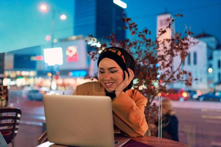 En el encantador ambiente de un paisaje urbano nocturno, una chica vestida de hiyab absorta en su computadora portátil crea una imagen fascinante, que encarna la fusión de la tecnología, el empoderamiento y la vibrante ciudad.