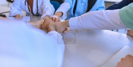 Un grupo de médicos y una enfermera médica unen sus manos en una mesa, mostrando el trabajo en equipo inquebrantable y la solidaridad que impulsa sus esfuerzos colectivos en el campo de la salud. 