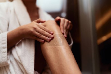 Una mujer se entrega a su rutina de cuidado personal, mimando su piel después de un baño relajante mediante la aplicación de crema hidratante en sus piernas, abrazando la esencia de la belleza y el bienestar. 