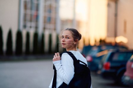 A beautiful blonde teenager returns home from school during the sunset, carrying her school backpack, in a serene and peaceful urban setting. 