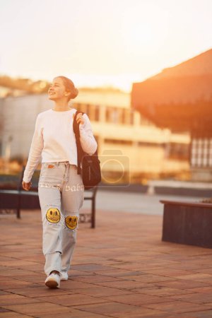 Eine schöne blonde Teenagerin kommt bei Sonnenuntergang mit ihrem Schulrucksack von der Schule nach Hause, in einer ruhigen und friedlichen städtischen Umgebung. 