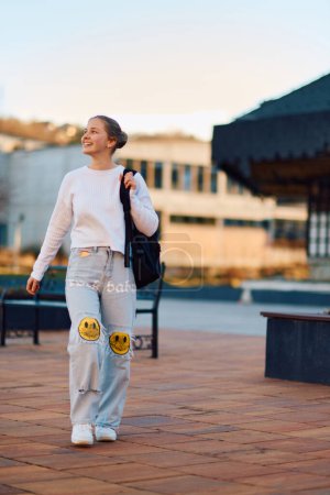 Eine schöne blonde Teenagerin kommt bei Sonnenuntergang mit ihrem Schulrucksack von der Schule nach Hause, in einer ruhigen und friedlichen städtischen Umgebung. 