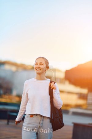 Une belle adolescente blonde rentre de l "école au coucher du soleil, portant son sac à dos scolaire, dans un cadre urbain serein et paisible. 