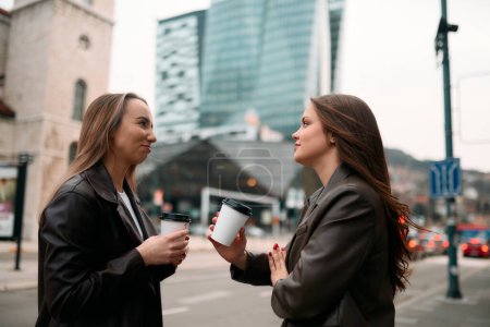 Two businesswomen take a break outside their company, enjoying a moment of relaxation as they hold cups of coffee in their hands. 