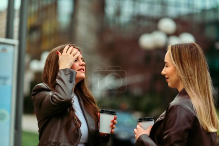 Two professional women stand outside their office, sharing a coffee and engaging in a friendly conversation during their break.