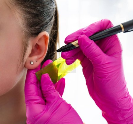 Kosmetikerinnen in rosa sterilen Handschuhen halten Lineal und Stift in der Hand, um die Stelle eines weiblichen Ohrs zu messen, um den Ohrring zu durchstechen. Hochwertiges Foto