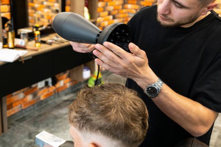 The picture of a professional barber is with a hair diffuser that he uses to make curly hair of a blond and freckled boy. The boy wants to have curly and beautiful hair. High quality photo