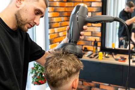 The picture of a professional barber is with a hair diffuser that he uses to make curly hair of a blond and freckled boy. The boy wants to have curly and beautiful hair. High quality photo
