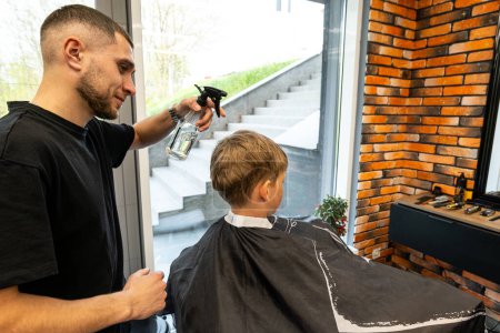 Photo for A boy sits in a barbershop while he is sprayed with water to start a haircut. A master in a barbershop sprays water on a boy who is sitting and waiting for his haircut. . High quality photo - Royalty Free Image