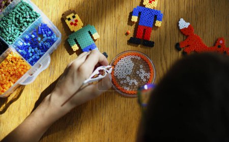 Téléchargez les photos : Boy's hand holding tweezers with a bead, creating art toy from fused colorful beads (also perler beads or iron beads). Toy developing children's imagination. Children education concept. From above. - en image libre de droit