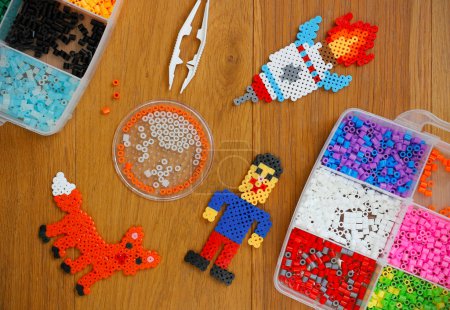 Téléchargez les photos : Colorful iron beads (perler or fused beads) in a box, art toys created from them and tweezers on wooden background. Toy developing children's imagination. Concept of children's creativity. Top view - en image libre de droit