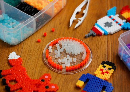 Téléchargez les photos : Colorful iron beads (perler beads or fused beads) in a box, art toys created from them and white tweezers on wooden background. Toy developing children's imagination. Concept of children's creativity. - en image libre de droit