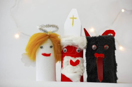 Nikolaus, Engel und Teufel von einem Kind aus Toilettenpapierrollen auf weißem Hintergrund mit Weihnachtsbeleuchtung gebastelt. Einfache diy kreative Weihnachtsidee. Umweltfreundliche Recycling-Dekoration.