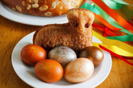 Osterstillleben mit einem kleinen Lammkuchen, bemalten Eiern, dem traditionellen tschechischen Ostergebäck "mazanec" und einer Osterpeitsche mit bunten Bändern im Hintergrund. Traditionelle tschechische Ostersymbole