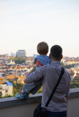 Photo for A Caucasian father wearing a checkered shirt hugs his 3-year-old son and together they look at the panorama of Prague in the evening summer sun. Shot from behind. Father's Day concept with copy space. - Royalty Free Image