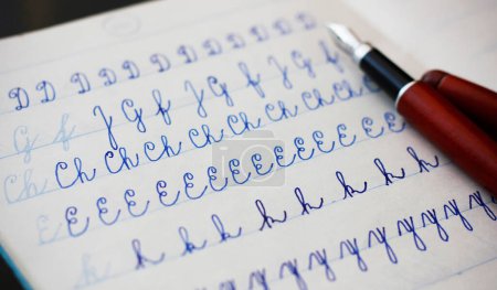 Close-up of uppercase and lowercase letters written by a child in cursive with a fountain pen with blue ink in a lined notebook, a burgundy fountain pen in the background. Concept for school education and handwritten documents.
