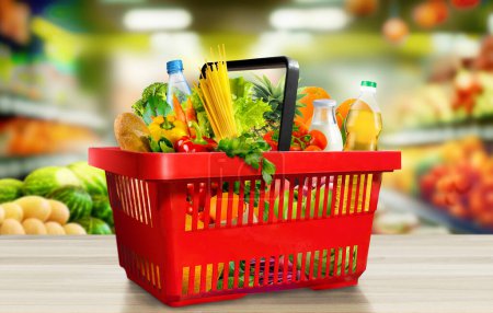 Food and groceries in red shopping basket on wood table on blurred suppermarket aisle background