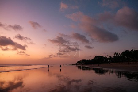 Téléchargez les photos : Sous l'éclat enchanteur du coucher de soleil orange, les amateurs de plage se délectent de l'ambiance sereine de Kuta Beach. À gauche, les eaux calmes reflètent les tons chauds du ciel - en image libre de droit