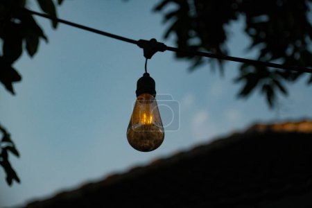Téléchargez les photos : Plongez-vous dans l'ambiance enchanteresse de la nuit alors qu'une ampoule tamisée suspendue sur une ficelle métallique projette une douce lueur contre l'obscurité veloutée. - en image libre de droit