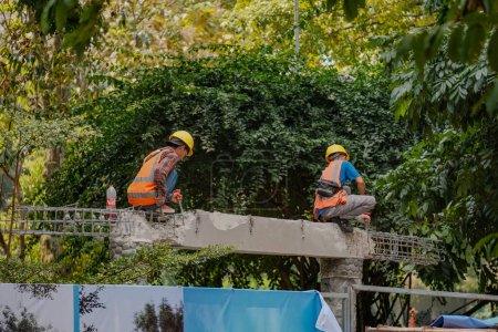 Téléchargez les photos : Travailleurs indonésiens de la construction intensément concentrés sur un pieu de béton, martelant avec précision sur un fond bordée d'arbres, mettant en valeur l'artisanat qualifié en cours. - en image libre de droit