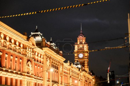 The iconic Flinders Street Station illuminated by vibrant orange lighting against the night sky, with its distinctive clocktower casting a timeless silhouette.