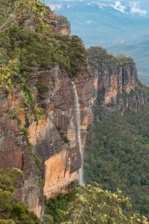 A tranquil scene in the Blue Mountains of Australia, where a slender waterfall cascades gently amidst the rugged beauty of the landscape