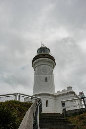 A striking view of Byron Bay Lighthouse from its base, framed against a backdrop of cloudy skies, showcasing its enduring presence