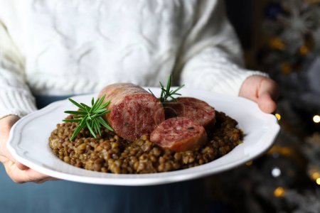 Photo for Italian food concept. Hands holding white plate with Italian large pork sausage cotechino served with lentils. Christmas decorations. Christmas holiday. Close-up. - Royalty Free Image