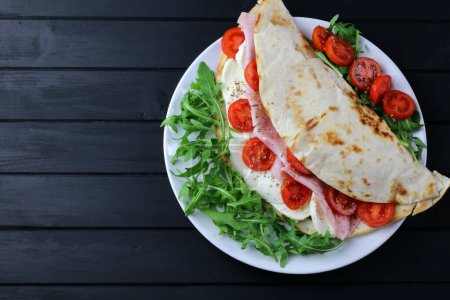 Photo for Italian piadina with ham, mozzarella, rocked salad and tomatoes isolated on wooden black background. Overhead view. Copy space. - Royalty Free Image