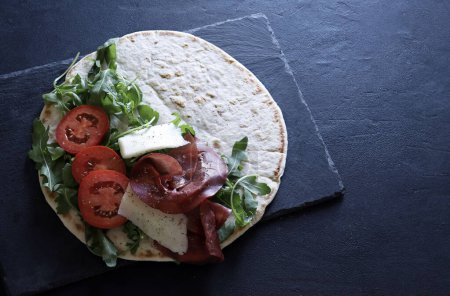 Photo for Traditional Italian piadina with ham, cheese, rocked salad and tomatoes isolated on black background. Italian food street. Overhead view. Copy space. - Royalty Free Image