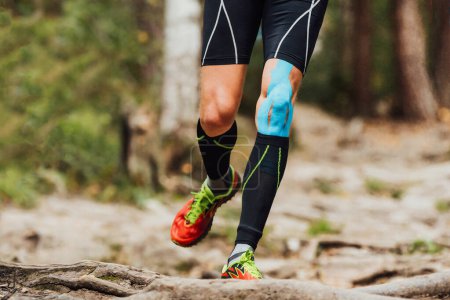 Téléchargez les photos : Jambes coureur masculin avec bande kinesio sur genou courir marathon de sentier forestier, chaussettes de compression noires et kinésiotaping bleu - en image libre de droit