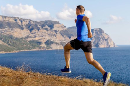 Foto de Corredor masculino corriendo sendero de montaña a lo largo de la orilla del océano, ropa deportiva camisa azul y pantalones cortos negros - Imagen libre de derechos