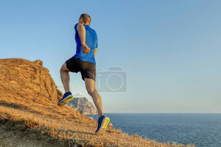 Photo for Back male runner run uphill mountain trail in blue sky background, summer jogging along coast at sunset - Royalty Free Image